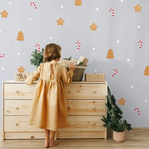 Jouluteemainen seinäkoriste, jossa on piparkakkuja joulukuusien ja tähtien muodossa sekä candy canes, ja pieni mekkoon pukeutunut tyttö seisoo lipaston edessä.
