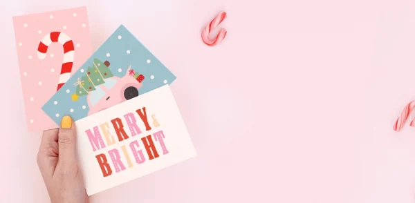 Käsi, joka pitää värikkäitä syntymäpäiväkortteja jouluteemalla ja teksteillä “Merry Bright” ja “Ho Ho Ho” lahjapaketin päällä.