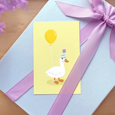 Keltainen lasten syntymäpäiväkortti, jossa on juhlahattuinen hanhi ja keltainen ilmapallo, asetettuna violetilla rusetilla koristellun lahjan päälle.