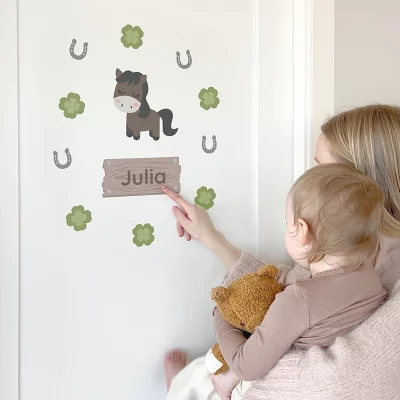 Äiti ja lapsi ihailevat lastenhuoneen seinätarraa, jossa on hevosen kuva ja nimeä Julia. Tarra on koristeltu hevosenkengillä ja apiloilla, luoden henkilökohtaisen ja leikkisän ilmeen lapsen huoneeseen.