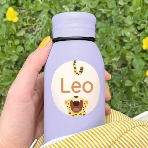 Vaaleanvioletti juomapullo, johon on kiinnitetty suuri nimilappu tekstillä Leo, leikkisä leopardi-aihe, ulkona vihreällä taustalla.