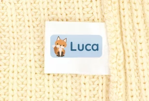 Vaatteen pesumerkintälappuun kiinnitetty sininen nimitarra, jossa ketun kuva ja teksti "Luca".