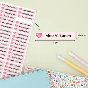 Suloisia nimilappuja, joissa on Aino Virtanen -nimikirjoitus sydänkuvioineen. Nimilaput ovat käytännöllisiä varusteita lasten tarvikkeisiin ja leikkeihin. Taustalla näkyy värikkäitä muistikirjoja ja kyniä.