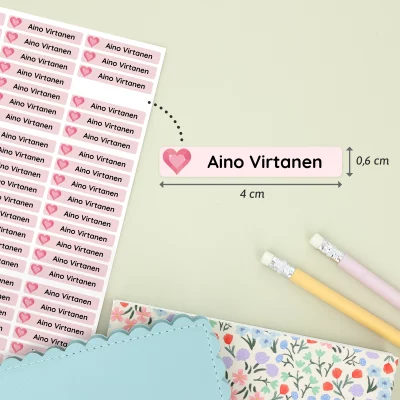 Suloisia nimilappuja, joissa on Aino Virtanen -nimikirjoitus sydänkuvioineen. Nimilaput ovat käytännöllisiä varusteita lasten tarvikkeisiin ja leikkeihin. Taustalla näkyy värikkäitä muistikirjoja ja kyniä.