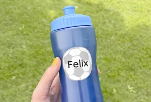 Sininen juomapullo, jossa on henkilökohtainen nimilappu Felix jalkapalloteemalla. Pullo on kädessä vihreällä nurmikolla, mikä tekee siitä täydellisen ulkoleikkeihin ja urheilutoimintaan.
