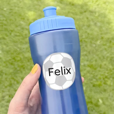 Sininen juomapullo, jossa on henkilökohtainen nimilappu Felix jalkapalloteemalla. Pullo on kädessä vihreällä nurmikolla, mikä tekee siitä täydellisen ulkoleikkeihin ja urheilutoimintaan.