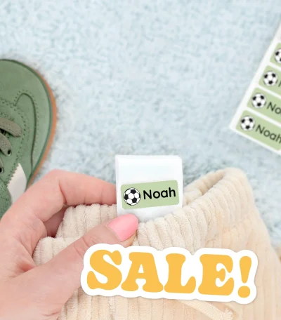 Nuoret kenkät, joissa on nimilappu Noah, ovat esillä pehmeällä taustalla. Kuvassa näkyy myös vaatteeseen liimattu nimi- ja jalkapallokuvioitu etiketti. Ideal apparel for kids, joissa on mahdollisuus personoida omilla nimillä.