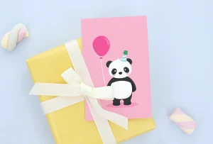Syntymäpäiväkortti, jossa on suloinen panda, joka pitää vaaleanpunaista ilmapalloa. Kortti on asetettu keltaisen lahjapaketin päälle, joka on koristeltu valkoisella nauhalla. Tämä kortti on täydellinen lisä syntymäpäivälahjoihin lapsille.