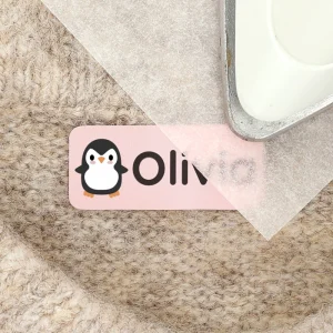 Nimiä etiketti, jossa on pingviini-kuvio ja nimi Olivia, asetettuna pehmeän taustatekstiilin päälle. Täydellinen valinta lasten vaatteiden ja tavaroiden merkitsemiseen.